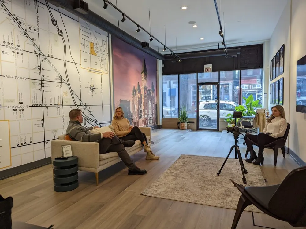 Real estate agents filming an interview-style video series in a Chicago storefront office, with a camera on a tripod